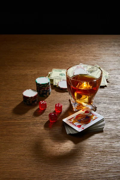 Vista ad angolo alto di vetro di cognac vicino alle banconote del dollaro, mazzo di carte, fiches da casinò e dadi sulla superficie isolata su nero — Foto stock