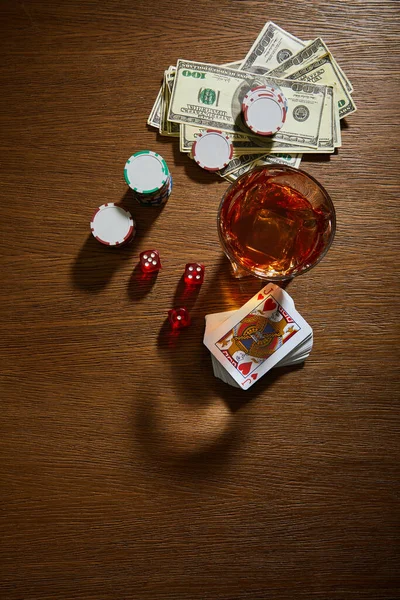 Vista superior de vidro de conhaque perto de notas de dólar, baralho de cartões, fichas de casino e dados em fundo de madeira — Fotografia de Stock