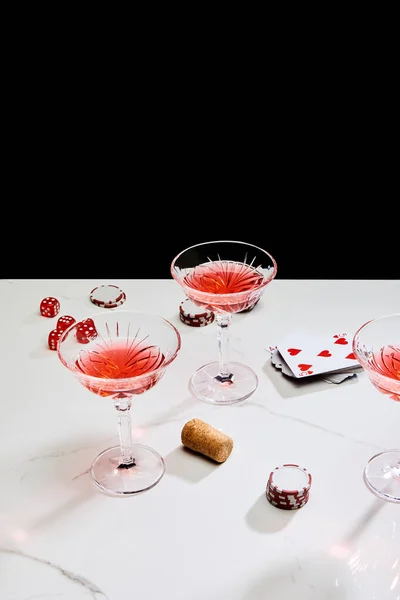 High angle view of glasses of cocktail, dice, cork, casino chips and playing cards on white surface isolated on black — Stock Photo