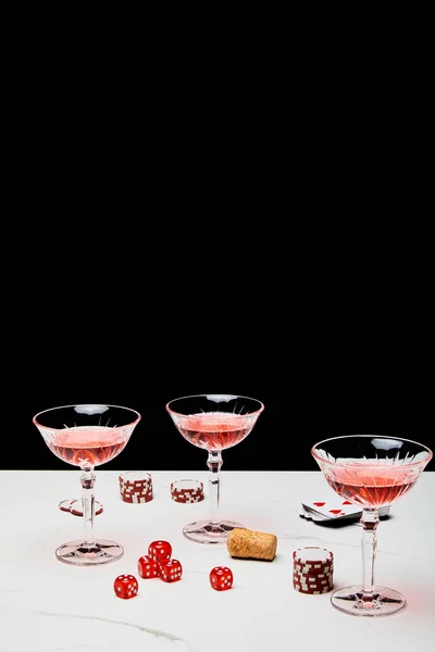 Glasses of cocktail, dice, cork, casino tokens and pack of cards on white surface isolated on black — Stock Photo
