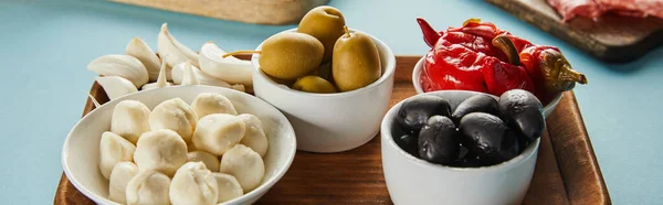 Olives, mozzarella and marinated chili peppers in bowls with garlic on board on blue, panoramic shot — Stock Photo