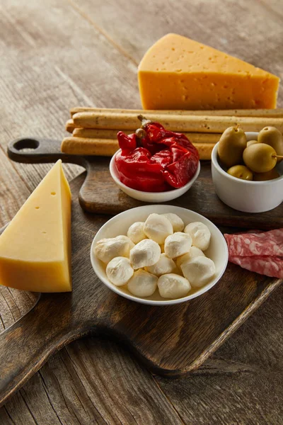 Vista de alto ângulo de tigelas com ingredientes antipasto perto de queijo, fatias de salame e paus de pão em placas em fundo de madeira — Fotografia de Stock