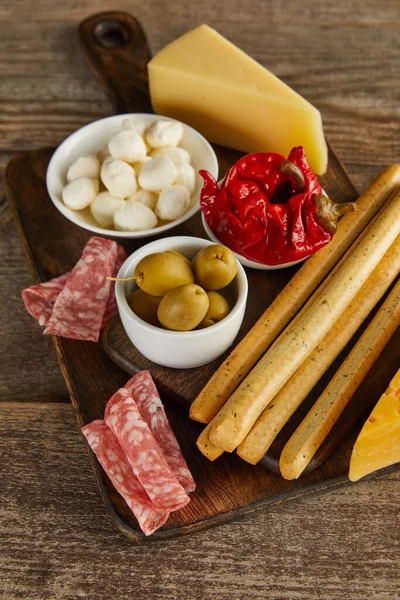 Vista de alto ângulo de tigelas com ingredientes antipasto com paus de pão, fatias de salame e queijo em placas em fundo de madeira — Fotografia de Stock