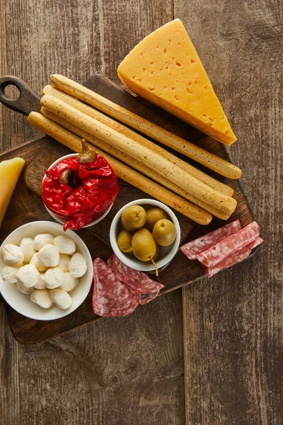 Vue du dessus des bols avec des ingrédients antipasto près de bâtonnets de pain, tranches de salami et fromage sur des planches sur fond en bois — Photo de stock