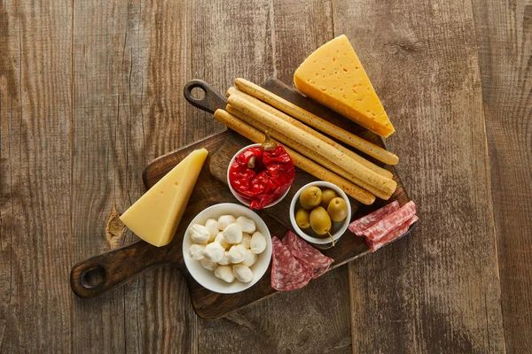 Top view of bowls with antipasto ingredients near breadsticks, salami slices and cheese on boards on wooden background — Stock Photo