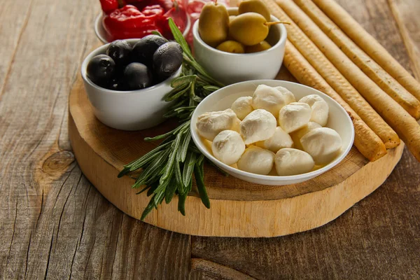 Round board with rosemary, breadsticks and bowls with antipasto ingredients on wooden background — Stock Photo