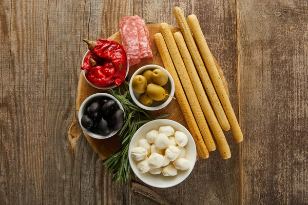 Vista dall'alto di grissini, fette di salame e rosmarino vicino a ciotole con ingredienti antipasto su tavola rotonda su fondo di legno — Foto stock