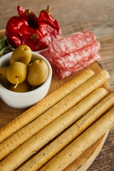 High angle view of breadsticks and salami slices near bowls with marinated chili peppers and olives on board on wooden background — Stock Photo