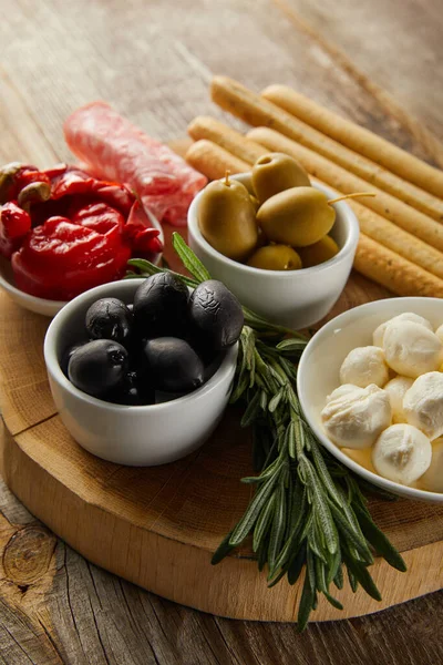 Selective focus of breadsticks, salami slices and rosemary near bowls with antipasto ingredients on board on wooden background — Stock Photo