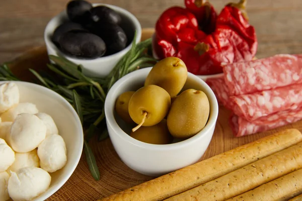 High angle view of bowls with olives, chili peppers and mozzarella near breadsticks, salami slices and greenery on board on wooden background — Stock Photo