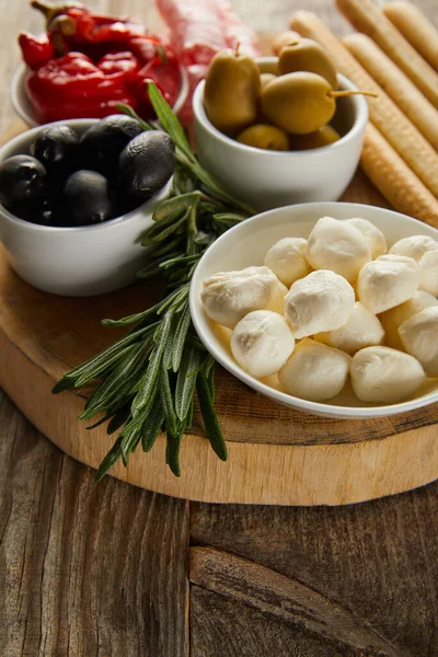 Selective focus of bowls with olives, chili peppers and mozzarella near breadsticks and greenery on board on wooden background — Stock Photo