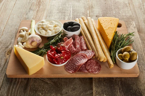 High angle view of board with cheese, breadsticks and antipasto ingredients on wooden background — Stock Photo