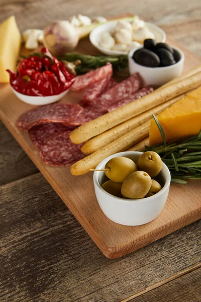 Selective focus of bowl with olives, breadsticks and antipasto ingredients on board on wooden background — Stock Photo