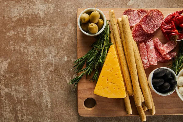 Top view of board with breadsticks, cheese and antipasto ingredients on brown background — Stock Photo