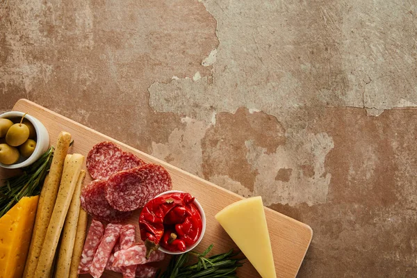 Top view of board with salami slices, cheese and antipasto ingredients on brown background — Stock Photo