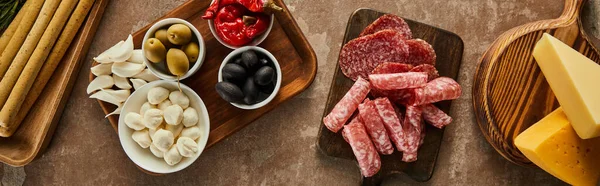 Top view of cheese, salami slices and antipasto ingredients on boards on brown background, panoramic shot — Stock Photo