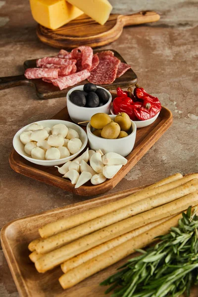 High angle view of boards with breadsticks, salami, rosemary, cheese and bowls with antipasto ingredients on brown — Stock Photo