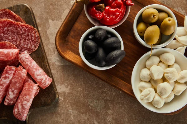 Top view of salami slices and bowls with olives, mozzarella and marinated chili peppers on boards on brown — Stock Photo