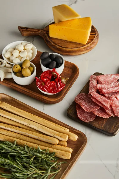 Vista en ángulo alto de tablas con palitos de pan, romero, rebanadas de salami, queso e ingredientes antipasto en blanco - foto de stock