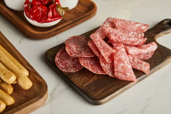 High angle view of salami slices, breadsticks and bowl with marinated chili peppers on boards on white — Stock Photo