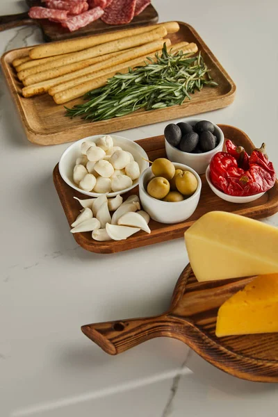 Vista ad alto angolo di formaggio, grissini, rosmarino, aglio e ciotole con ingredienti antipasto su tavole bianche — Foto stock