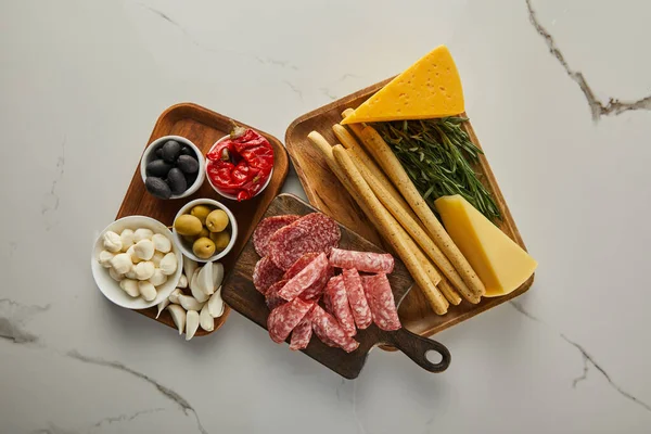 Top view of boards with salami slices, breadsticks, cheese, rosemary, garlic and bowls with antipasto ingredients on white — Stock Photo