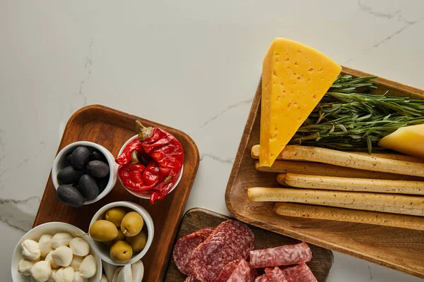 Top view of salami slices, breadsticks, cheese, greenery and bowls with antipasto ingredients on boards on white background — Stock Photo