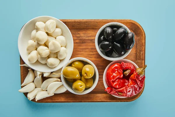 Top view of garlic and bowls with olives, mozzarella and marinated chili peppers on board on blue — Stock Photo