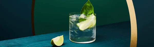 Vidrio con refrescante bebida con menta, hielo y rodaja de lima en la superficie de madera sobre fondo geométrico azul y verde - foto de stock