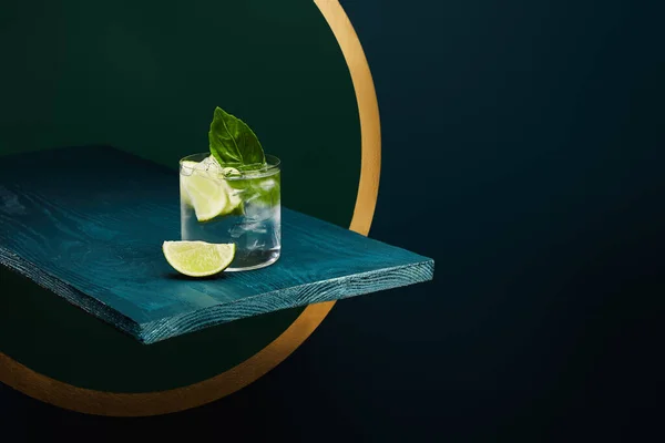 Vidrio con refrescante bebida con hielo, hoja de menta y rodaja de lima en la superficie de madera sobre fondo geométrico azul y verde - foto de stock