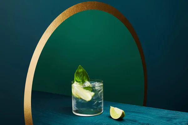 Vue grand angle du verre avec boisson rafraîchissante à la chaux, glaçons et feuille de menthe sur une surface en bois sur fond géométrique bleu et vert — Photo de stock