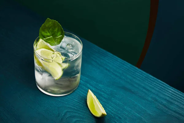 Hochwinkelblick auf altmodisches Glas mit Getränk und Limette auf blauer Holzoberfläche auf grünem und blauem geometrischem Hintergrund — Stockfoto