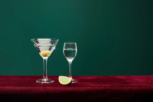 Shot glass and cocktail glass with vermouth, lime slice and whole olive on toothpick on velour surface isolated on green — Stock Photo