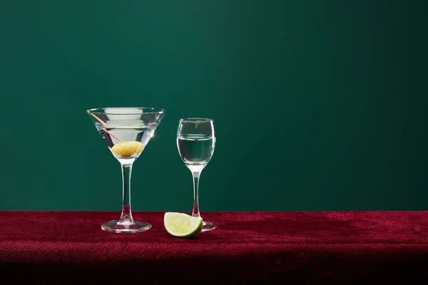 Vaso de chupito y copa de cóctel con vermut, rodaja de lima y aceituna entera en palillo de dientes en la superficie de terciopelo aislado en verde - foto de stock