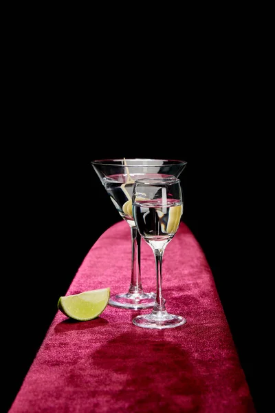 Рюмку и коктейльный бокал с вермутом, ломтиком лайма и целыми оливками на зубочистке на розовой велюровой поверхности, изолированной на черной — стоковое фото