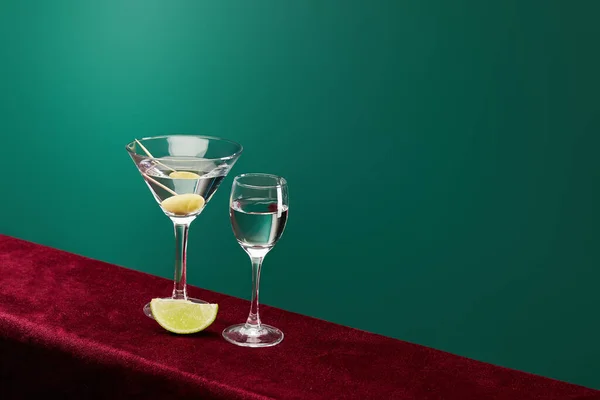 Vista ad alto angolo di bicchierino e bicchiere da cocktail con vermut, fetta di lime e olive intere su stuzzicadenti su superficie di velluto rosso isolata su nero — Foto stock