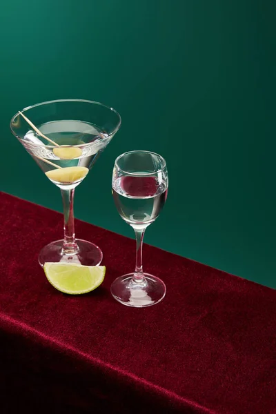 Hochwinkelaufnahme von Schnaps- und Cocktailglas mit Wermut, Limettenscheibe und ganzen Oliven auf Zahnstocher auf roter Velouroberfläche isoliert auf grünem Grund — Stockfoto