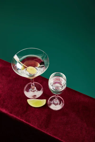 High angle view of shot glass and cocktail glass with vermouth, lime slice and whole olive on toothpick on velour surface isolated on green — Stock Photo