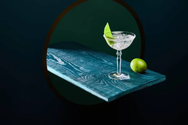 Hochwinkelaufnahme von Cocktailglas mit Minzblatt und ganzer Limette auf blauer Holzoberfläche auf geometrischem Hintergrund — Stockfoto
