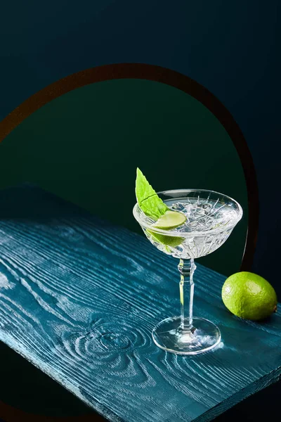 High angle view of cocktail glass with mint leaf and whole lime on blue wooden surface on geometric background with circle — Stock Photo