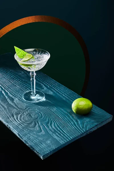 High angle view of cocktail glass with mint leaf and whole lime on blue wooden surface on geometric background with golden circle — Stock Photo