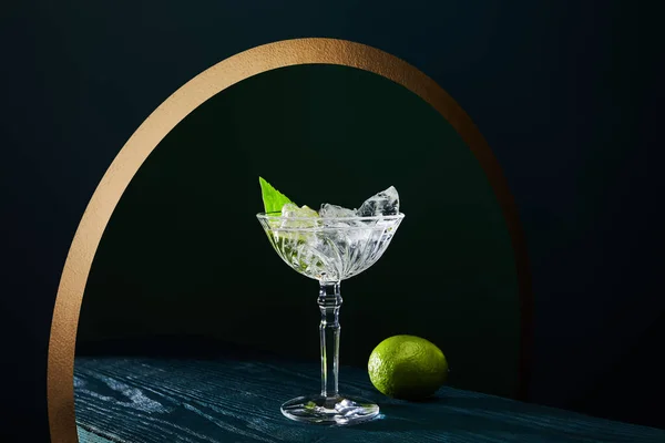 Cocktail glass with mint leaf and whole lime on blue wooden surface on geometric background with golden circle — Stock Photo