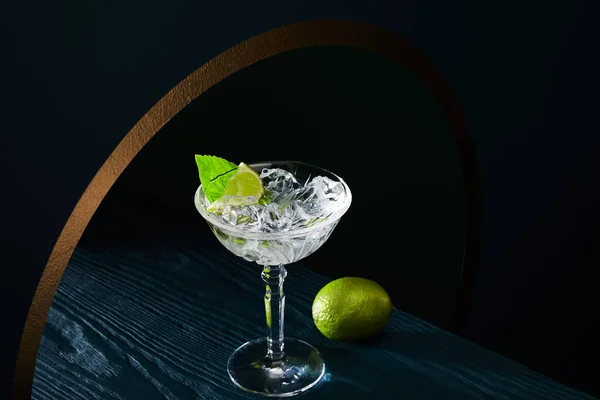 High angle view of cocktail glass with ice, mint leaf and whole lime on blue wooden surface on geometric background with golden circle — Stock Photo