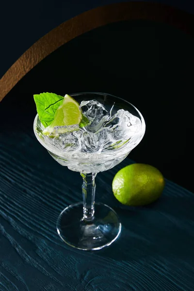 Alto angolo di vista del bicchiere da cocktail con ghiaccio, foglia di menta e tiglio intero su sfondo di legno blu — Foto stock