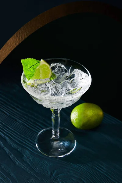 High angle view of cocktail glass with ice, mint leaf and whole lime on blue wooden background — Stock Photo