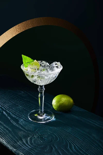 High angle view of cocktail glass with ice, mint leaf and whole lime on blue wooden background — Stock Photo