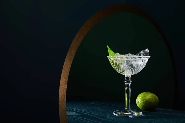 Cocktail glass with ice, mint leaf and whole lime on blue wooden surface on background with golden circle — Stock Photo