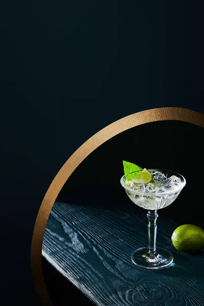 High angle view of cocktail glass with ice, mint leaf and whole lime on blue wooden surface on background with golden circle — Stock Photo