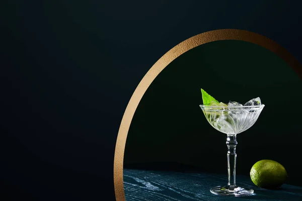 Cocktail glass with ice cubes, mint leaf and whole lime on blue wooden surface on geometric background with golden circle — Stock Photo