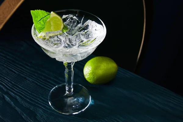 Hochwinkelblick auf Cocktailglas mit Eiswürfeln, Minzblatt und ganzer Limette auf blauer Holzoberfläche auf geometrischem Hintergrund — Stockfoto